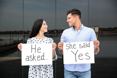 Photo of Lovely couple holding posters with text HE ASKED... SHE SAID YES after engagement outdoors