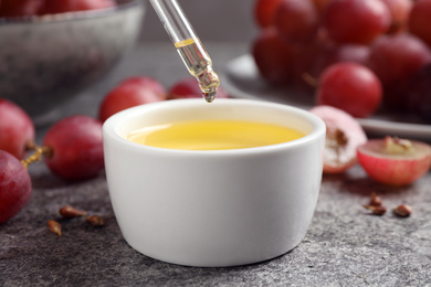 Photo of Dripping natural grape seed oil into bowl on grey table. Organic cosmetic