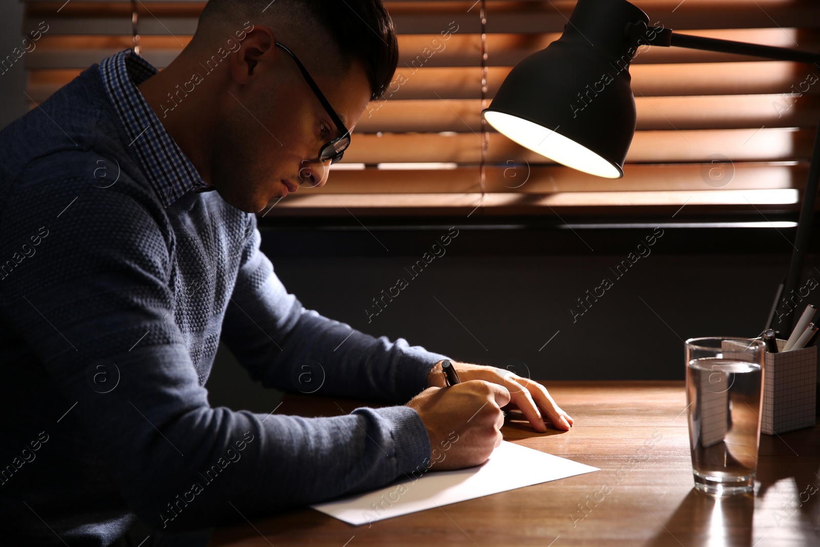 Photo of Man writing letter at wooden table in dark room