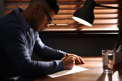 Man writing letter at wooden table in dark room