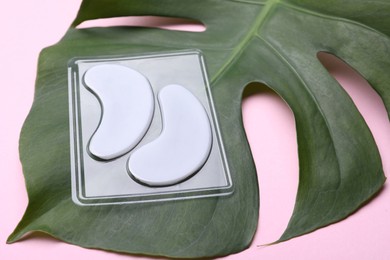 Photo of Package with under eye patches and green leaf on light pink background, closeup. Cosmetic product