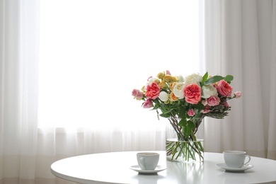 Vase with beautiful flower bouquet and cups of coffee on table in room. Space for text