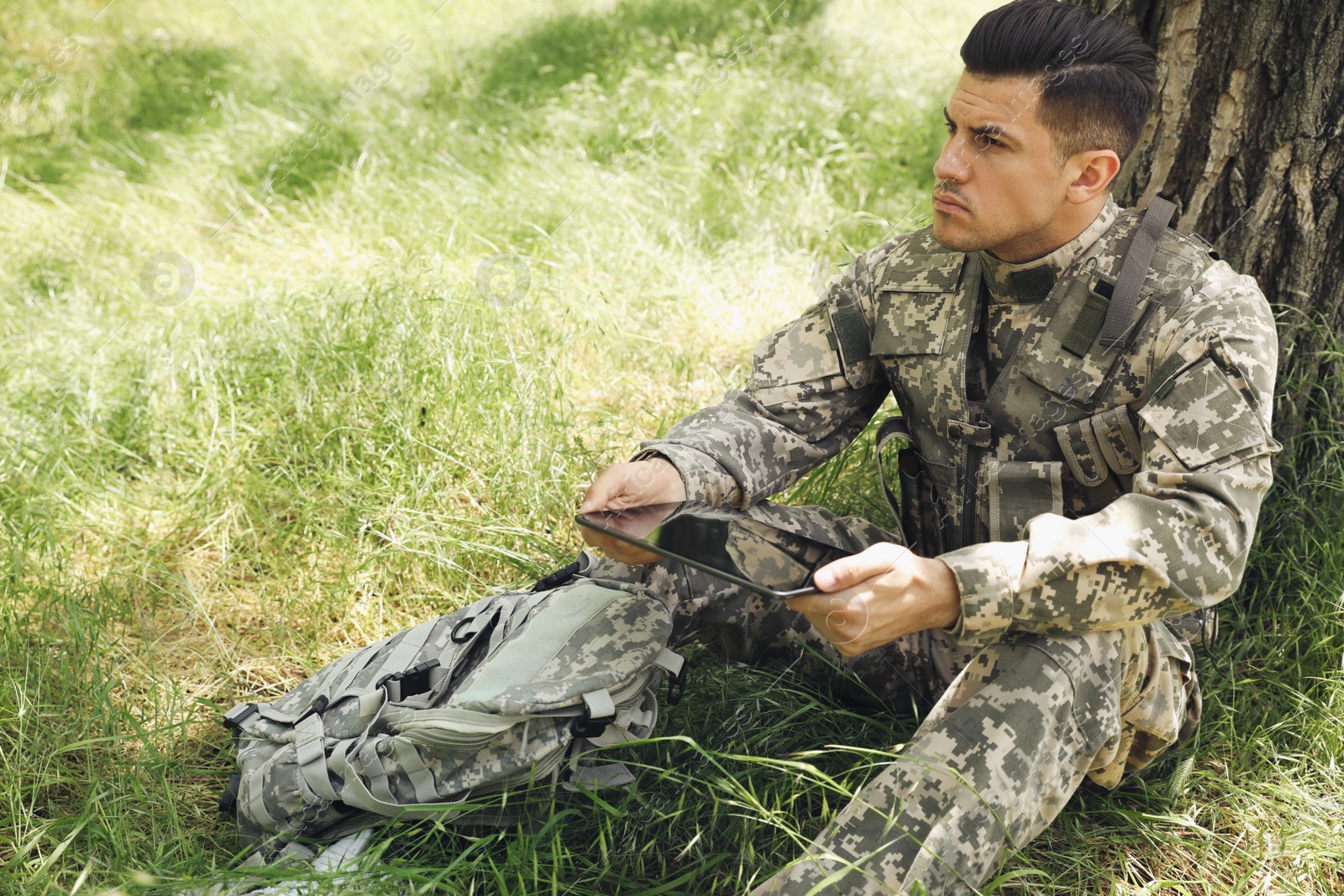 Photo of Soldier with backpack using tablet near tree in forest