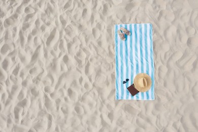 Striped beach towel with straw hat, book, sunglasses and slippers on sand, aerial view. Space for text