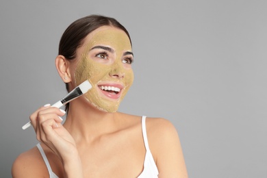 Photo of Young woman applying clay mask on her face against grey background. Space for text