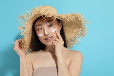 Beautiful young woman in straw hat with sun protection cream on her face against light blue background, space for text