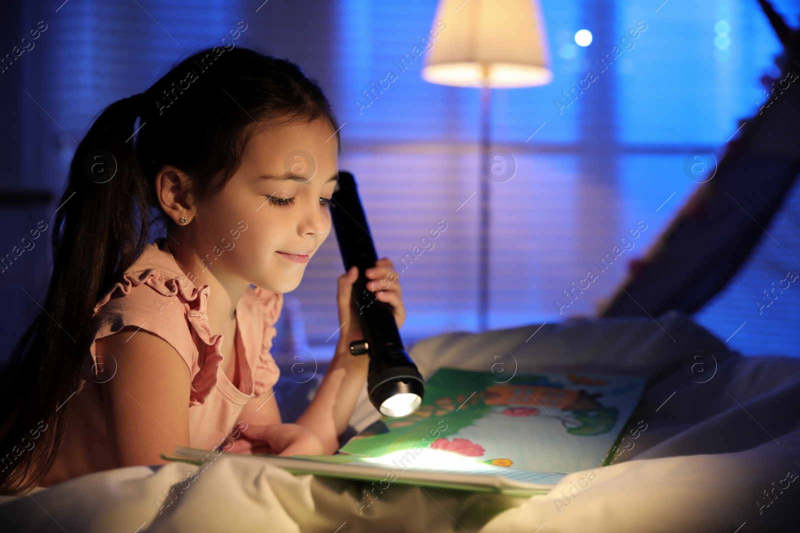 Photo of Little girl with flashlight reading fairy tale in dark bedroom