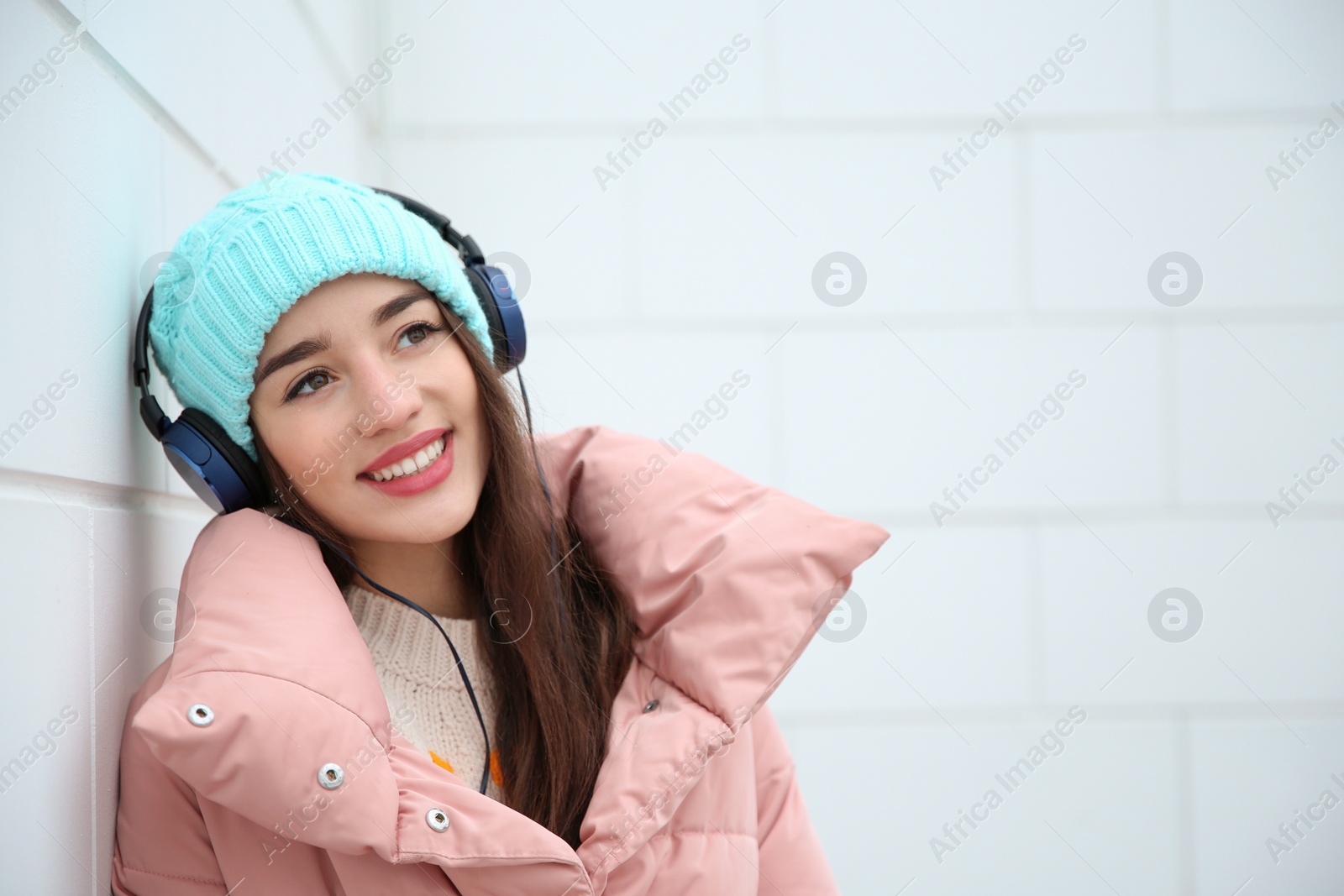 Photo of Beautiful young woman listening to music with headphones near light wall. Space for text