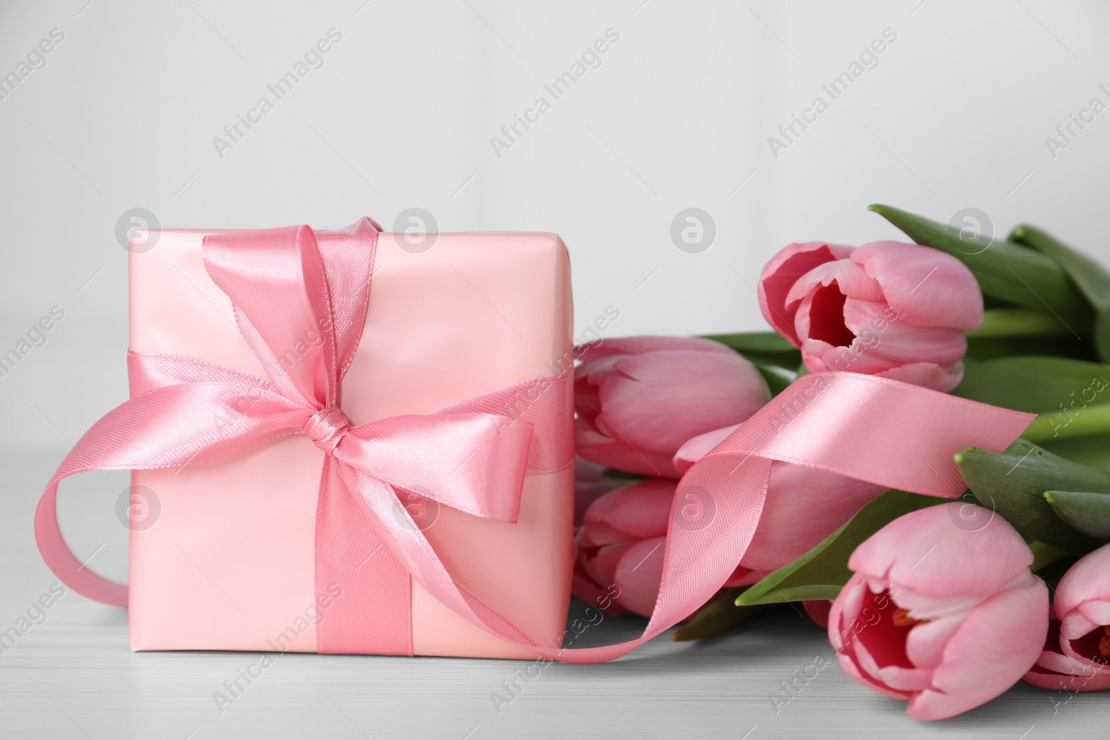 Photo of Beautiful gift box with bow and pink tulip flowers on light table, closeup