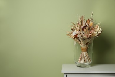 Photo of Beautiful dried flower bouquet in glass vase on white table near olive wall. Space for text