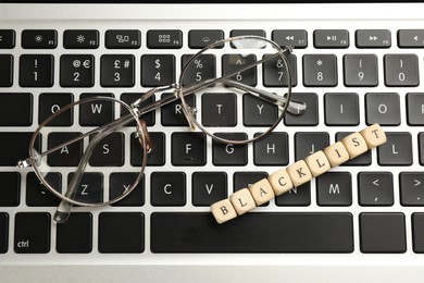 Photo of Wooden cubes with word Blacklist and glasses on laptop keyboard, top view