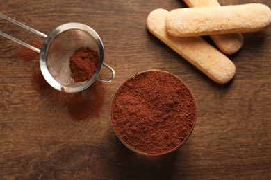 Photo of Delicious tiramisu in glass, biscuits and sieve with cocoa powder on wooden table, flat lay