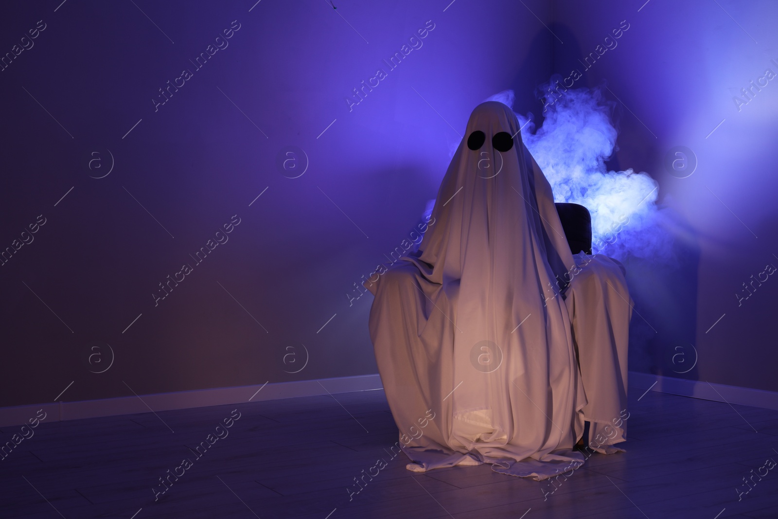 Photo of Creepy ghost. Woman covered with sheet sitting in armchair in blue light, space for text