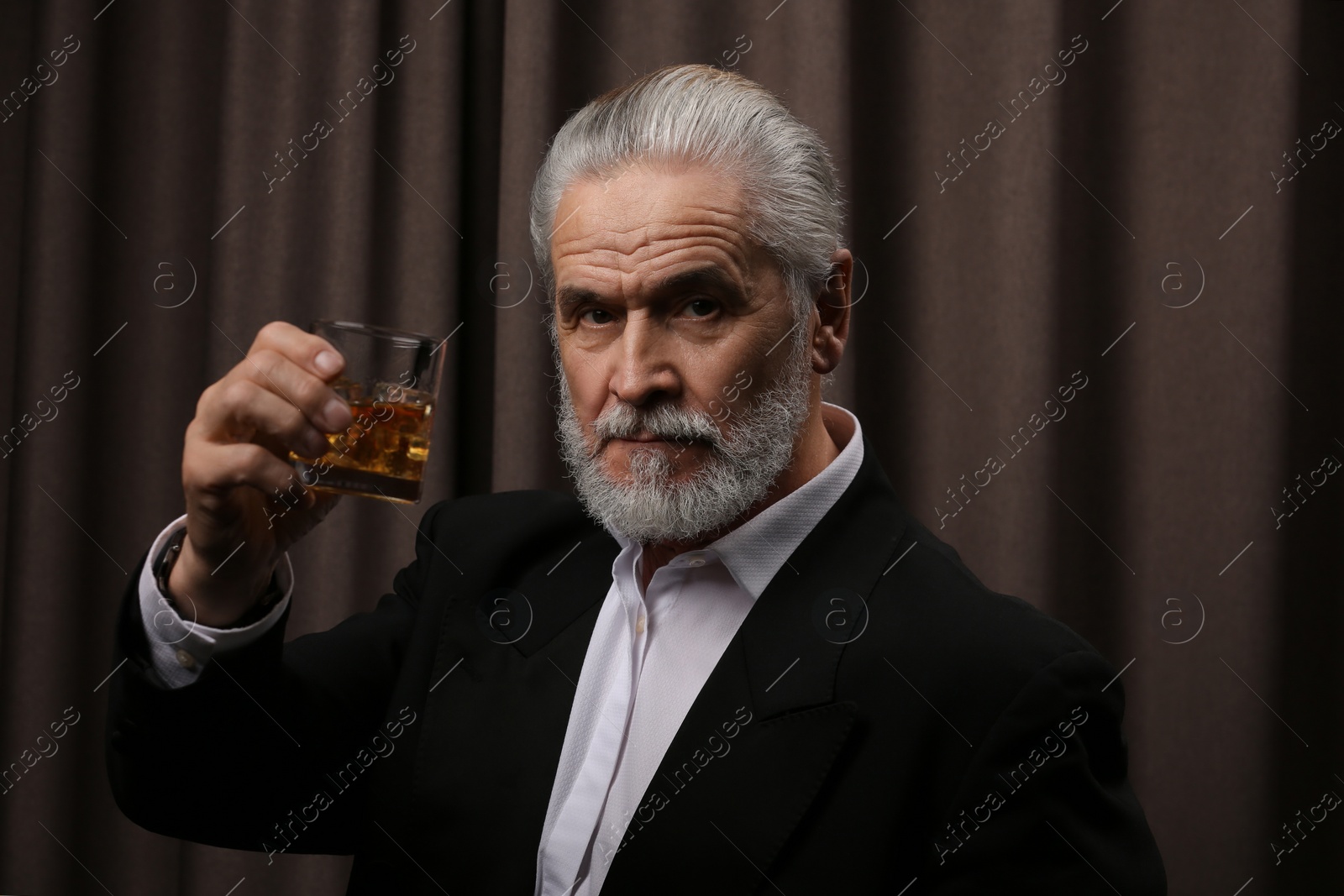 Photo of Senior man in formal suit holding glass of whiskey with ice cubes on brown background