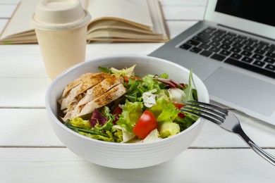 Photo of Bowl of tasty food, fork and laptop on white wooden table, closeup. Business lunch