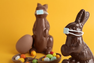 Photo of Chocolate bunnies with protective masks and candies on yellow background, closeup. Easter holiday during COVID-19 quarantine