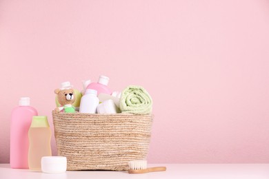 Photo of Baby cosmetic products, bath accessories and toy in wicker basket on white table against pink background. Space for text