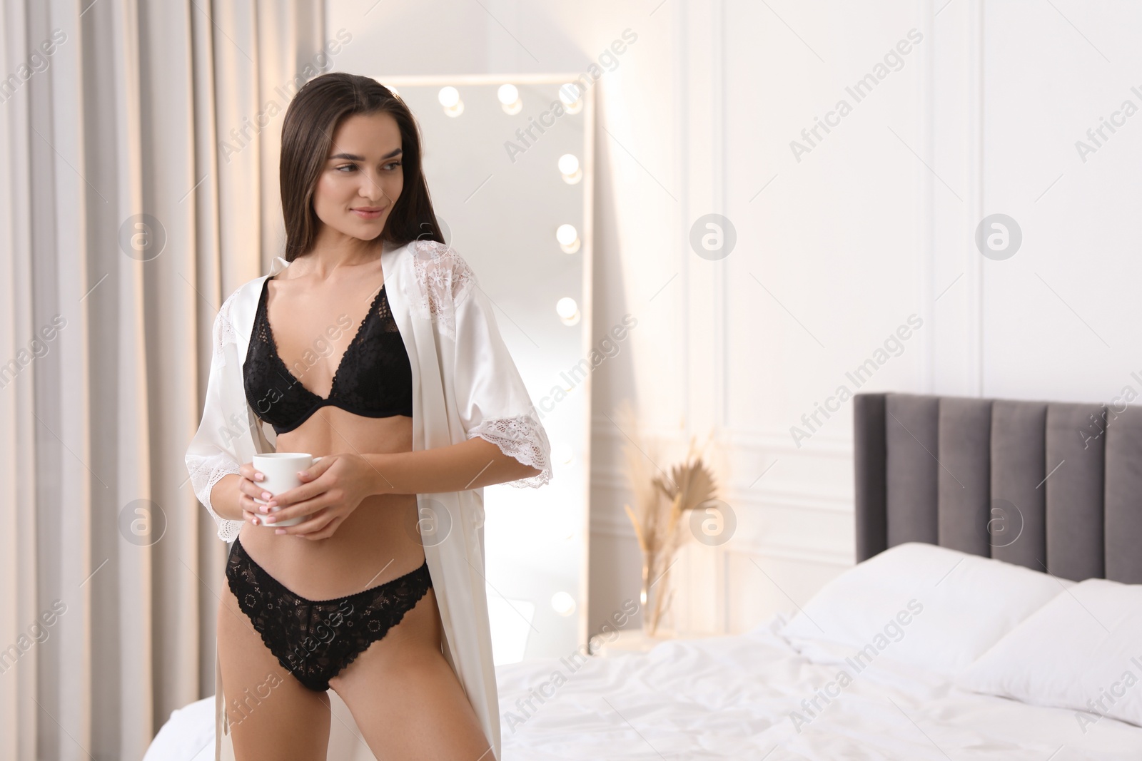 Photo of Young woman with cup of drink in elegant black underwear and robe indoors. Space for text