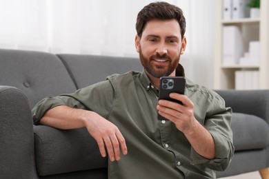 Photo of Smiling man using smartphone near sofa at home