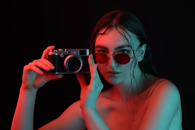 Photo of Beautiful woman with sunglasses and vintage camera posing in neon lights against black background