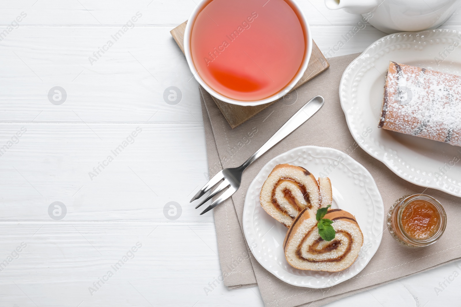 Photo of Tasty cake roll with jam and cup of tea on white wooden table, flat lay. Space for text