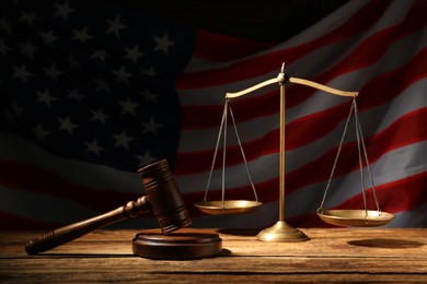 Photo of Scales of justice and judge's gavel on wooden table against American flag in darkness