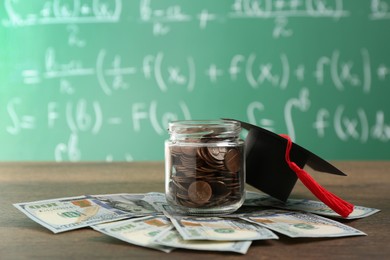 Photo of Scholarship concept. Glass jar with coins, graduation cap and dollar banknotes on wooden table, space for text