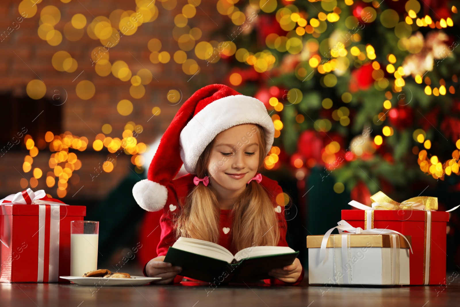 Photo of Little girl in Santa Claus cap reading book near Christmas tree indoors