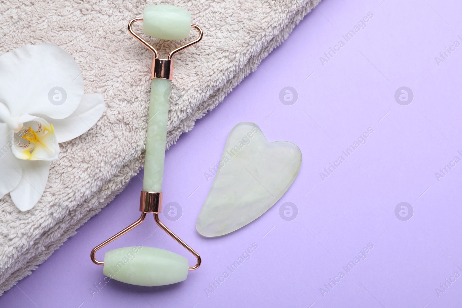 Photo of Gua sha stone, face roller, towel and orchid flower on violet background, flat lay. Space for text