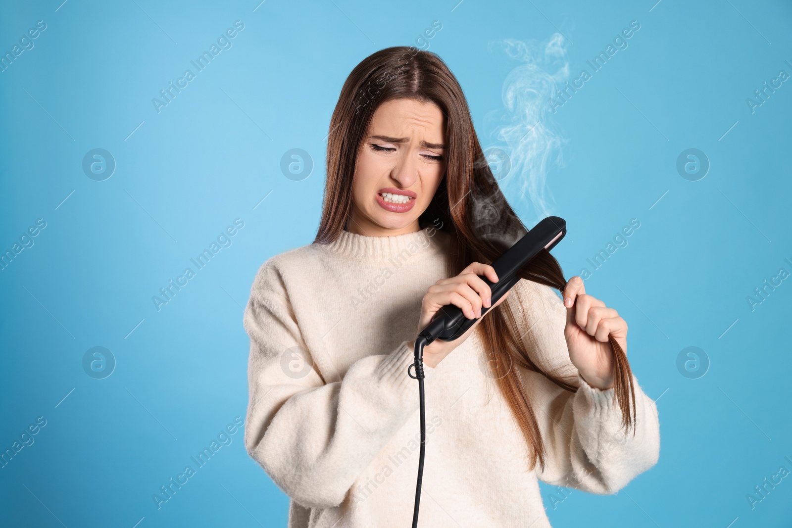 Photo of Stressed young woman with flattening iron on light blue background. Hair damage