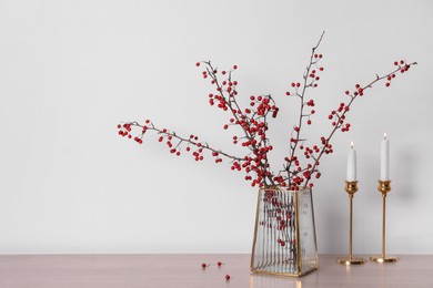 Photo of Hawthorn branches with red berries in vase and candles on wooden table, space for text