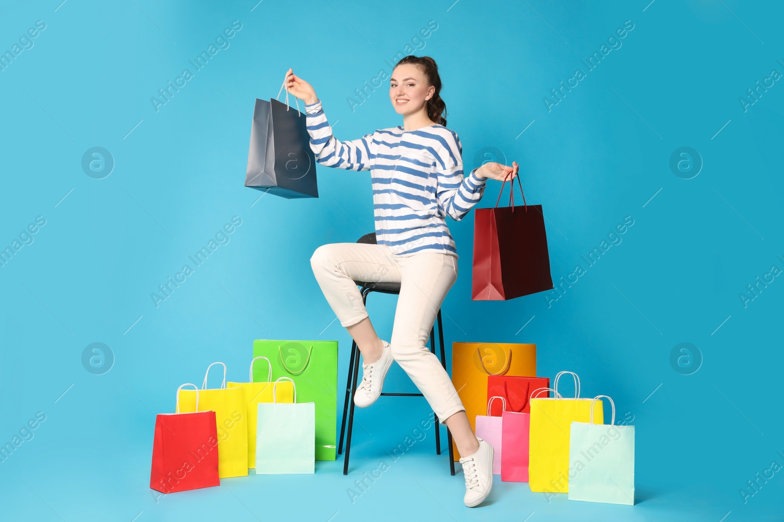 Photo of Happy woman holding colorful shopping bags on stool against light blue background
