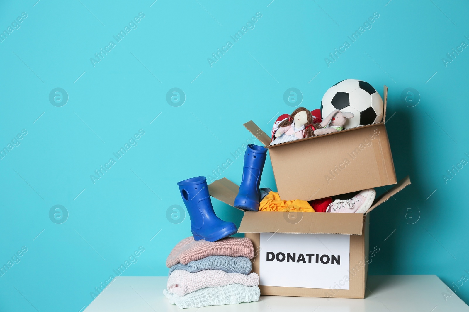 Photo of Donation boxes with toys, knitted clothes and shoes on table against color background. Space for text