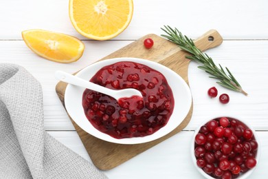 Photo of Tasty cranberry sauce in bowl, fresh berries, orange slices and rosemary on white wooden table, flat lay