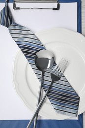 Photo of Plate with cutlery and tie on clipboard, top view. Business lunch concept