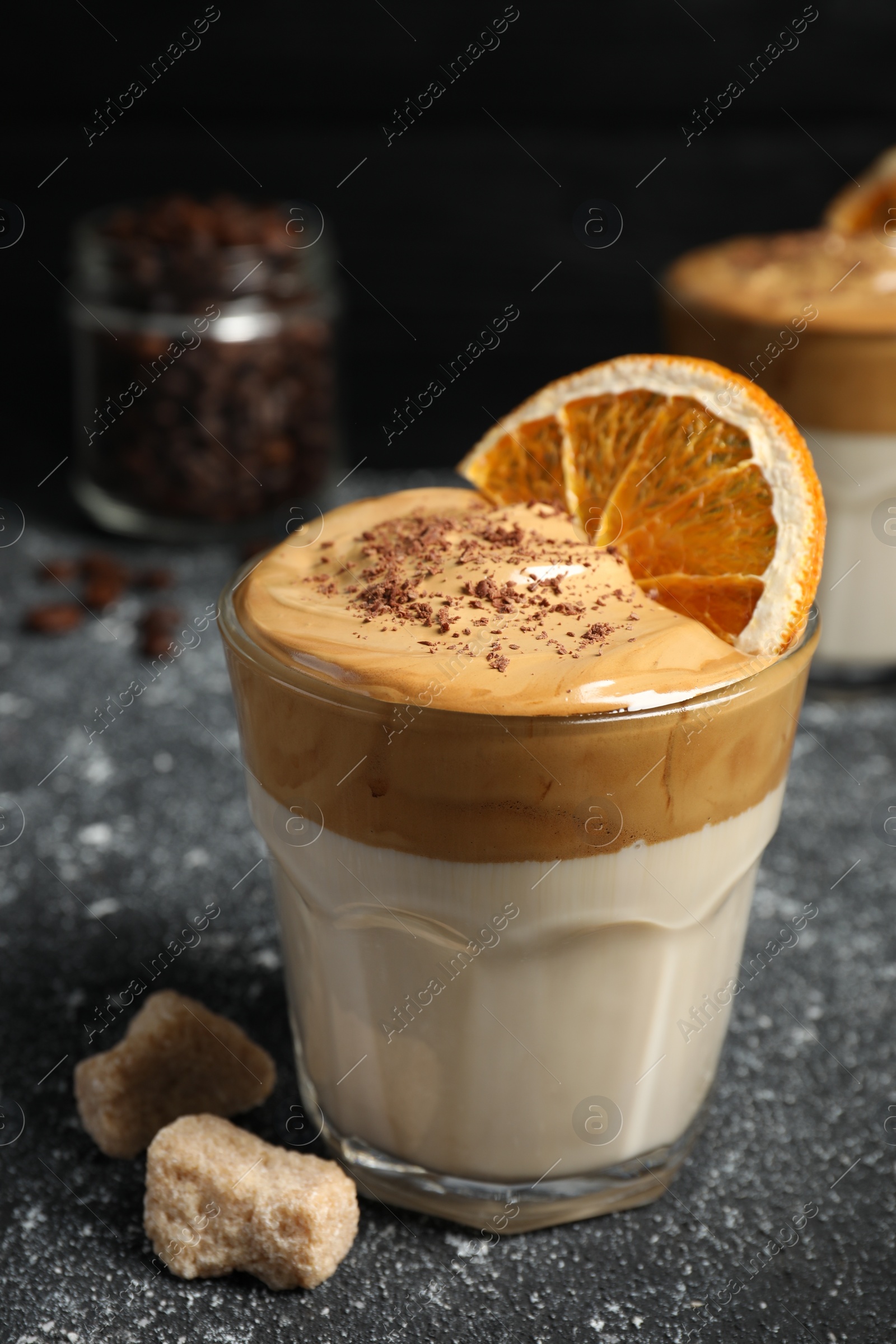 Photo of Glass of delicious dalgona coffee with dry orange, sugar and chocolate on grey textured table