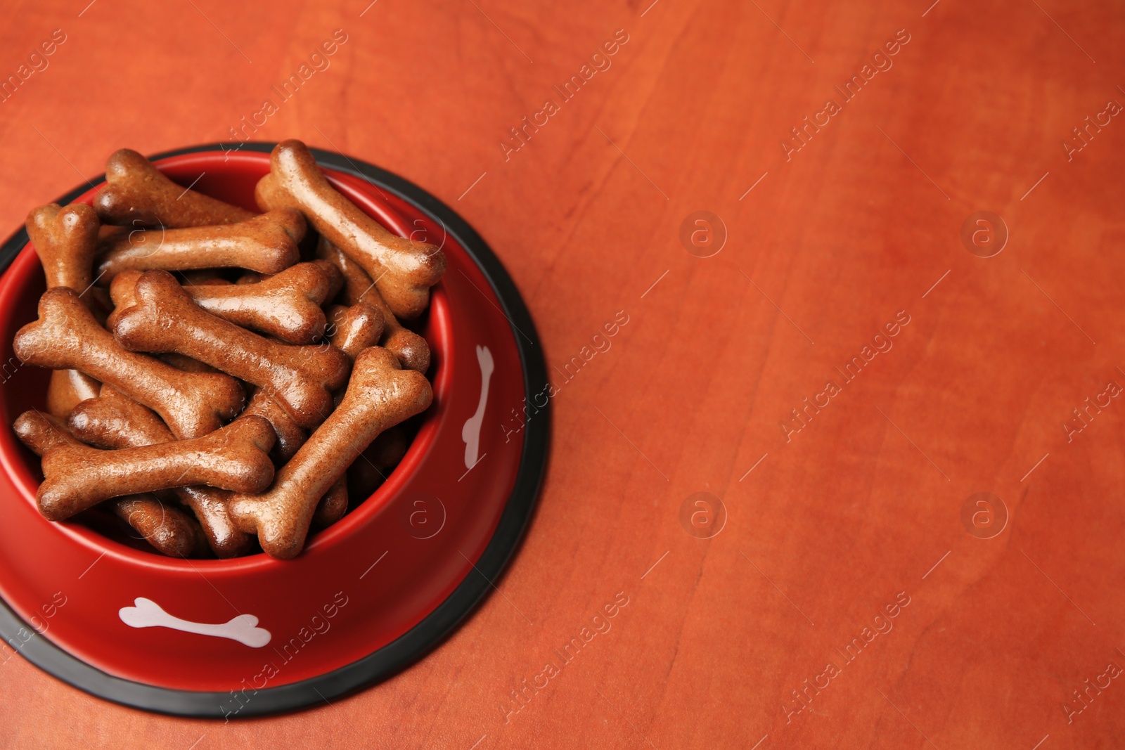 Photo of Red bowl with bone shaped dog cookies on wooden table, space for text