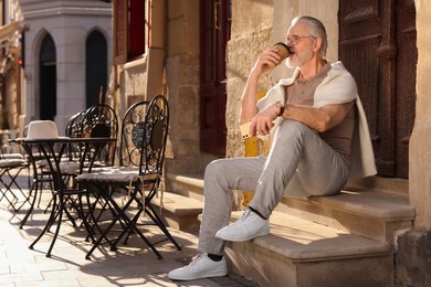 Photo of Handsome senior man sitting on doorstep with smartphone and drinking coffee outdoors, space for text