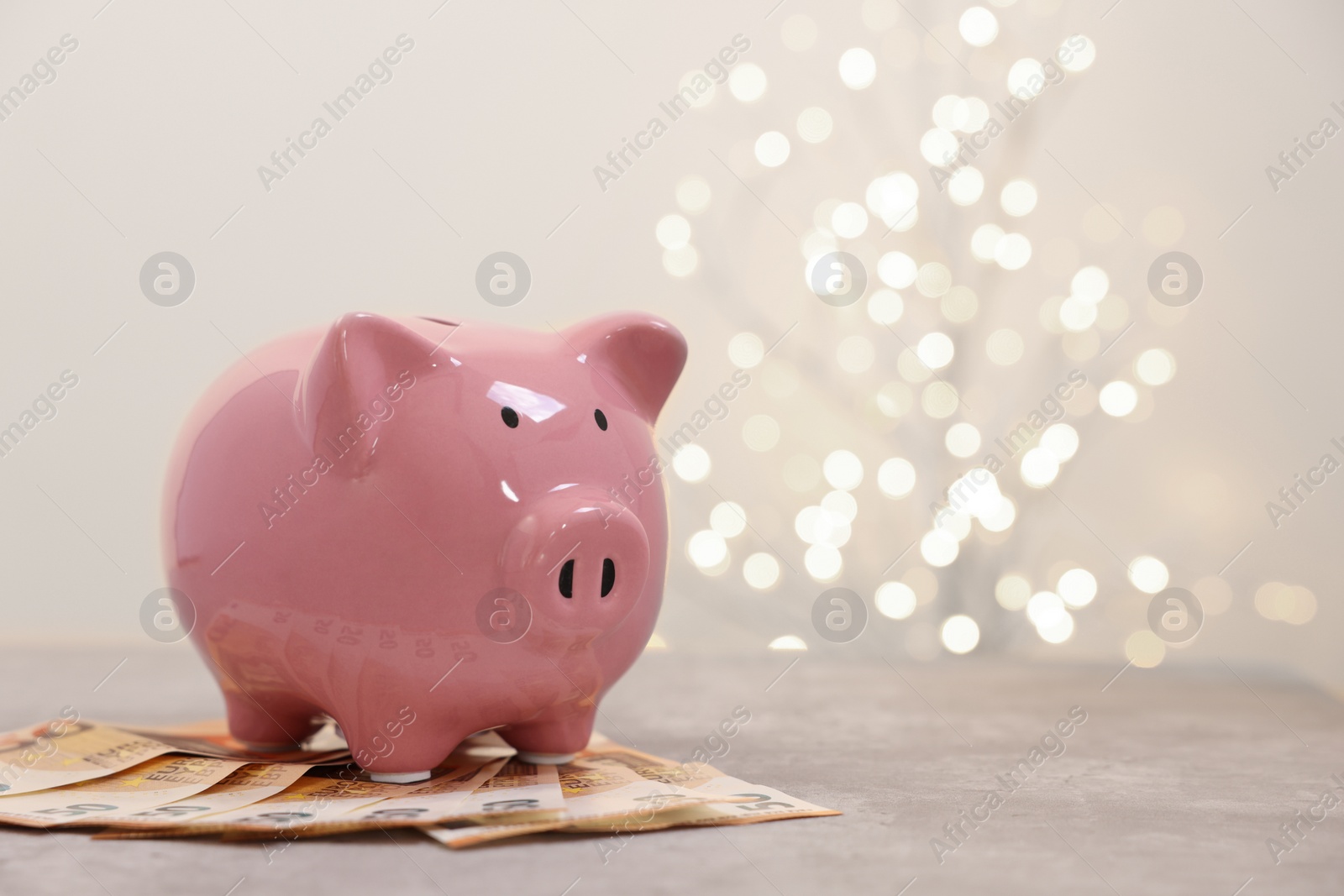 Photo of Piggy bank with euro banknotes on grey table against blurred lights, space for text