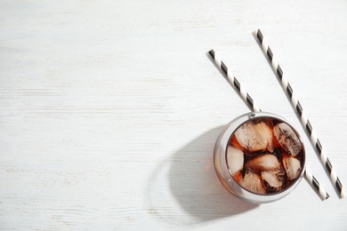 Glass of refreshing cola with ice cubes on white wooden background, top view. Space for text