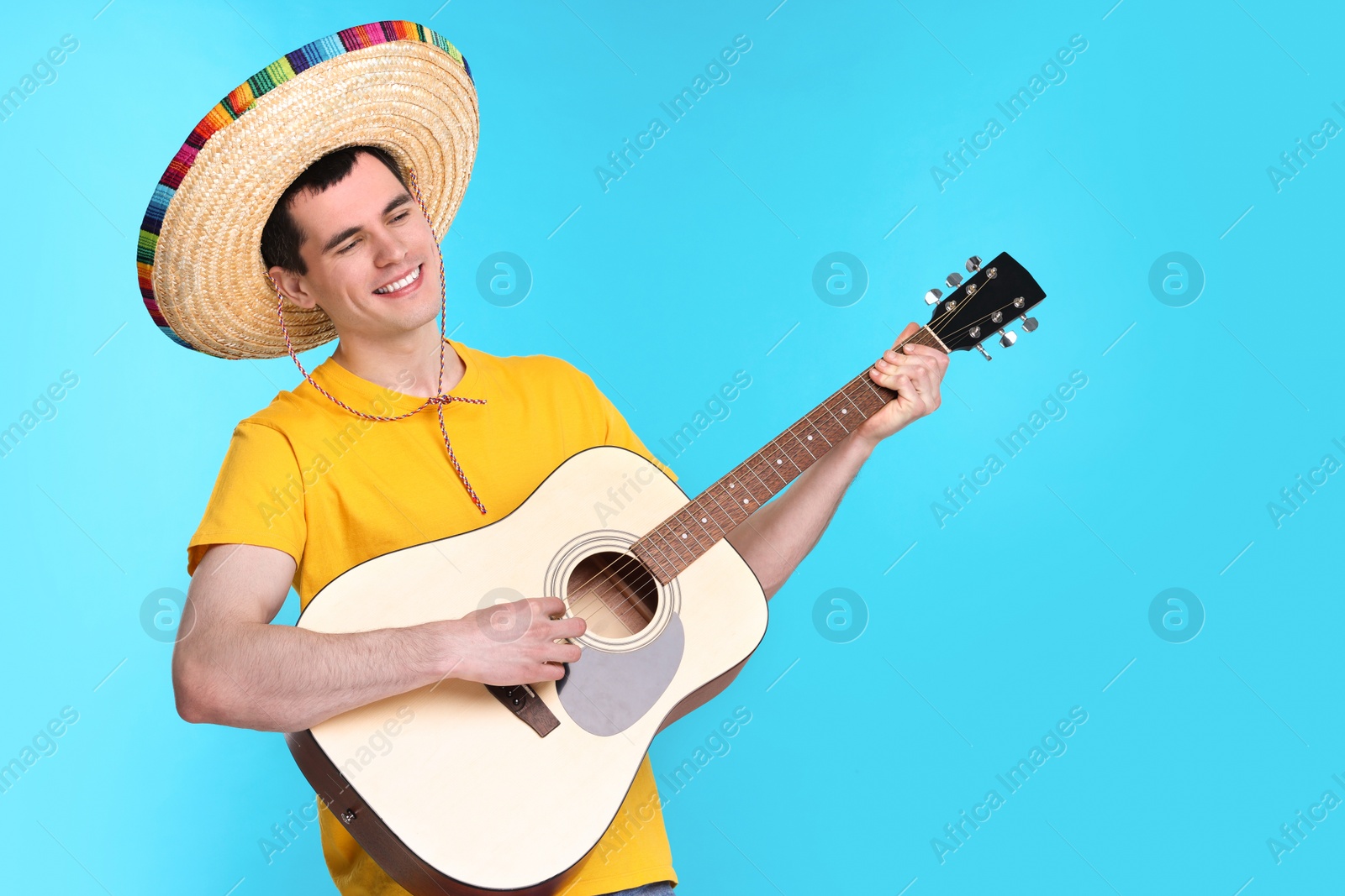 Photo of Young man in Mexican sombrero hat playing guitar on light blue background. Space for text