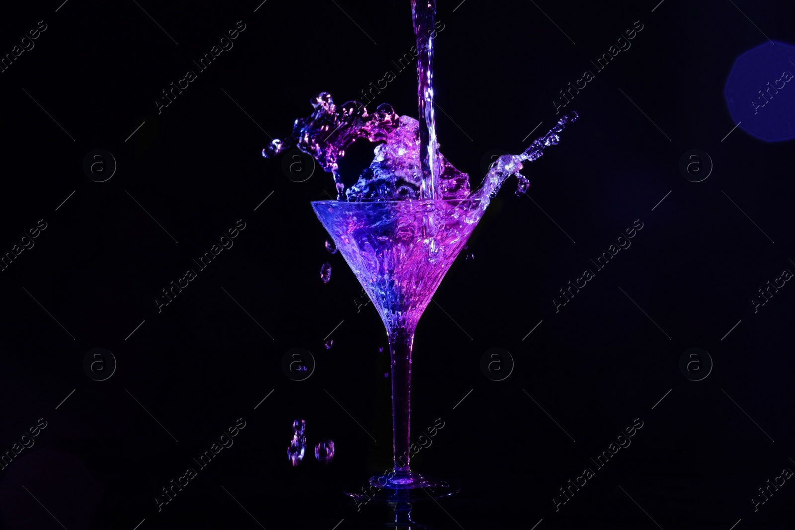 Photo of Pouring martini. Glass and splashes in neon lights on dark background, bokeh effect