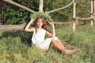 Cute little girl wearing wreath made of beautiful flowers near wooden fence outdoors on sunny day