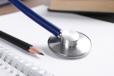 Photo of Student textbook, blank notebook, pencil and stethoscope on table, closeup. Medical education
