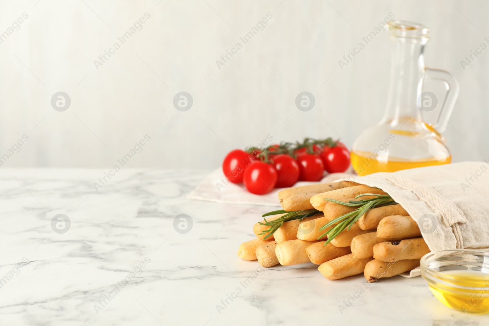 Photo of Delicious grissini sticks, oil, rosemary and tomatoes on white marble table. Space for text