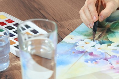 Photo of Woman painting flowers with watercolor at wooden table, closeup. Creative artwork