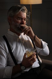 Photo of Bearded man with glass of whiskey smoking cigar in armchair indoors
