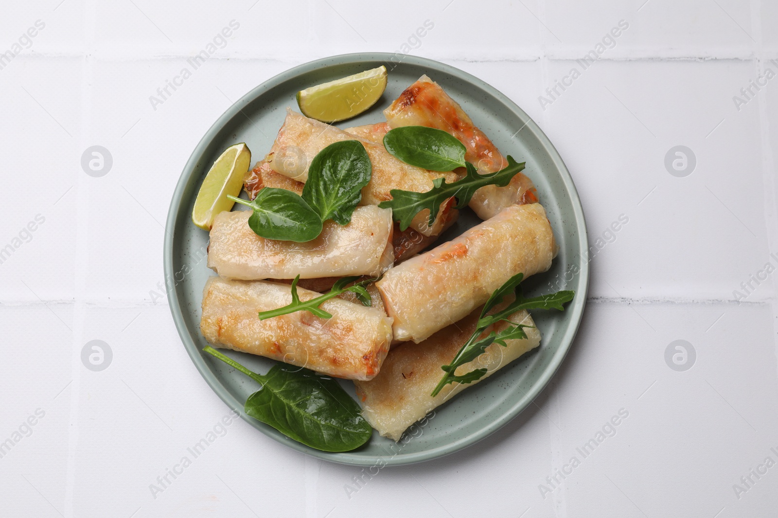Photo of Plate with tasty fried spring rolls, spinach, arugula and lime on white tiled table, top view