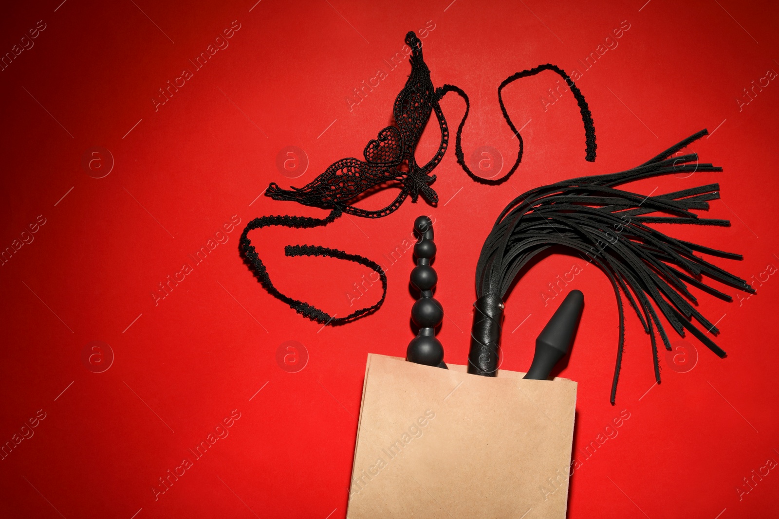 Photo of Paper shopping bag with different sex toys on red background, flat lay