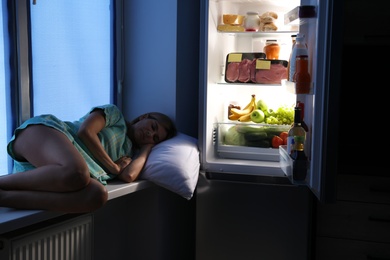 Woman sleeping on window sill near open refrigerator in kitchen at night